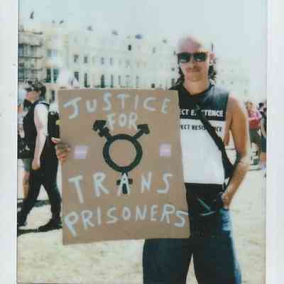 A person is holding a sign reading "Justice for Trans Prisoners" at an outdoor event.