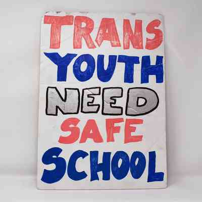 Hand painted, white, cardboard placard reads: "TRANS YOUTH NEED SAGE SCHOOL" in red, blue and silver
