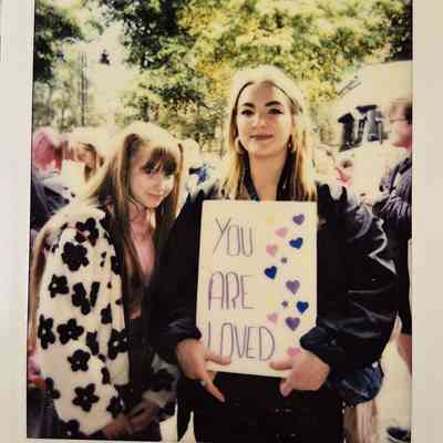 Two people stand outside, with one holding a sign that says, "You Are Loved."