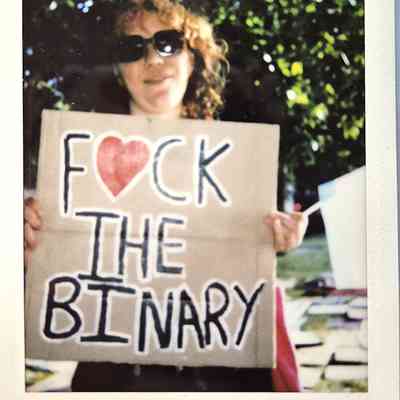A person wearing sunglasses holds a sign saying "F♥CK THE BINARY" at an outdoor event
