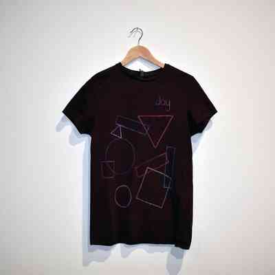 A burgundy t-shirt, printed with colored line drawings of geometric shapes and the word "Joy", on a wooden hangar.