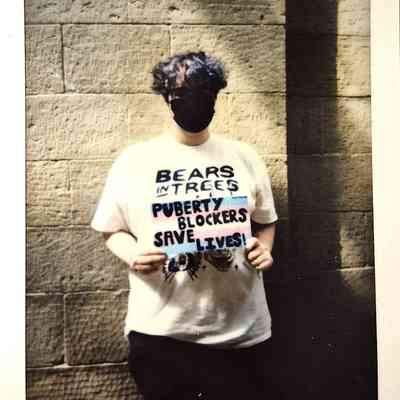 A person wearing a mask stands by a stone wall with a placard.
