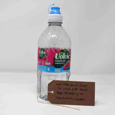 An empty plastic Volvic Touch of summer fruits water bottle. With a brown, handwritten luggage tag.