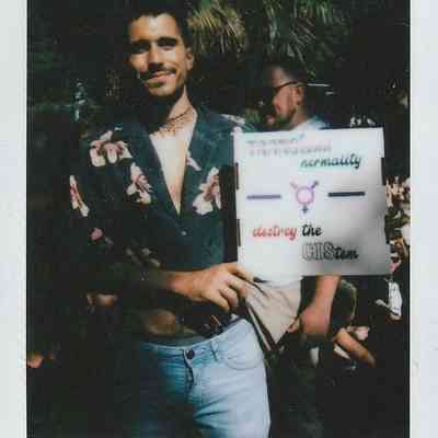 A smiling person in a floral shirt holds a sign which says something that is slightly blurry and hard to make out.