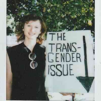 A person is standing outdoors, smiling beside a sign reading "THE TRANSGENDER ISSUE".