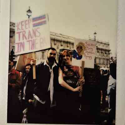 Two people hold signs which say "KEEP TRANS IN THE BAN" and "SASHAY AWAY CONVERSION THERAPY".