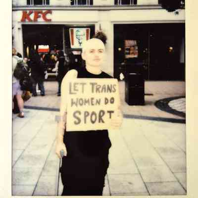 A person stands in front of a building, holding a sign supporting trans women's participation in sports.