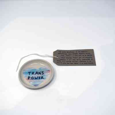 A small, white, ceramic dish illustrated with a blue, pink and white heart and the text "TRANS POWER" in black. With a brown, handwritten luggage tag.