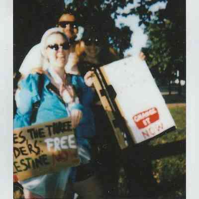 Three people are holding signs with messages during a protest event in a park setting.