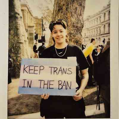A person holding a sign saying “KEEP TRANS IN THE BAN” is standing outdoors, smiling.