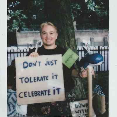 A person stands smiling, holding a sign that reads, "Don't just tolerate it, celebrate it".