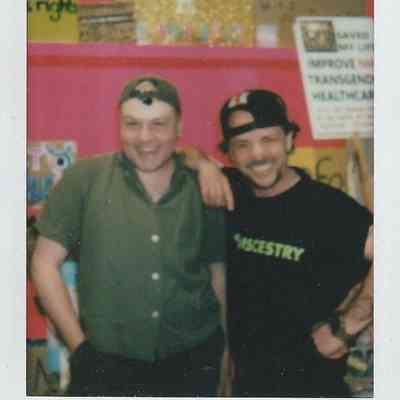 Two people stand smiling in front of a vibrant pink background with various posters.