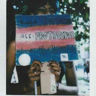 A person is holding a colorful sign with handwritten text, partially concealing their face.