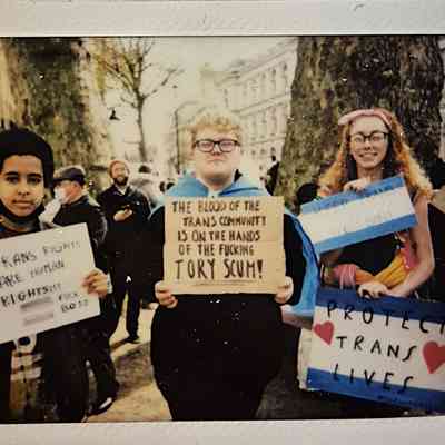 Three people are holding signs advocating for trans rights at a public protest. One says "TRANS RIGHTS ARE HUMAN RIGHTS FUCK BOJO", one says "THE BLOOD OF THE TRANS COMMUNITY IS ON THE HANDS OF THE FUCKING TORY SCUM" and ones says "PROTECT TRANS LIVES".