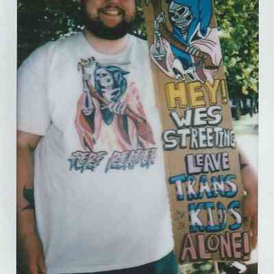 A person wearing sunglasses holds a sign reading "Leave Trans Kids Alone!" in a park setting.
