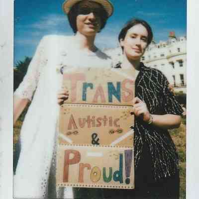 Two people stand together outdoors, holding a colorful sign reading "Trans Autistic & Proud".