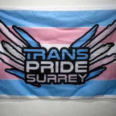 A blue, white and pink trans pride flag with "TRANS PRIDE SURREY" with 'wings' also in the trans pride colors.