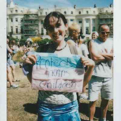 A person smiles while holding a sign reading "Protect Trans Children" at an outdoor gathering.