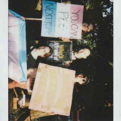 Three people hold signs advocating for transgender rights, standing outdoors with supportive messages.