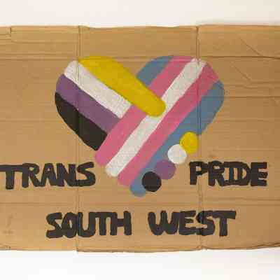 Hand painted brown cardboard placard with a stylised heart coloured in trans and non-binary colours and text "Trans Pride South West"