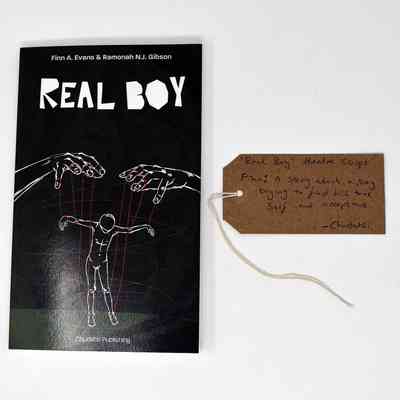 A black book which is called 'Real Boy' and has an illustration of a person being moved like a puppet. There is also a brown luggage tag with writing on it.