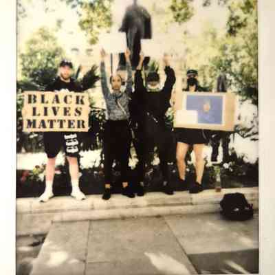 A diverse group stands with protest signs, supporting the Black Lives Matter movement, in a sunny park setting.