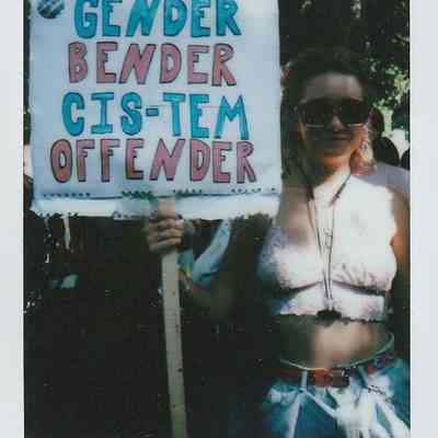A person holding a sign reads "Gender Bender Cis-tem Offender" at an outdoor event.