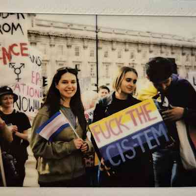 A group of people are standing at a protest, holding signs and smiling together in solidarity. The signs say 'FUCK THE CIS-TEM" and "RESISTANCE".