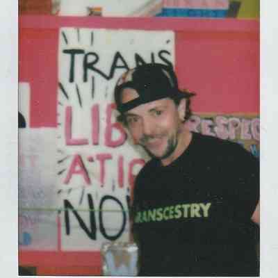 A person wearing a "TRANSCESTRY" shirt stands smiling in front of colorful posters.