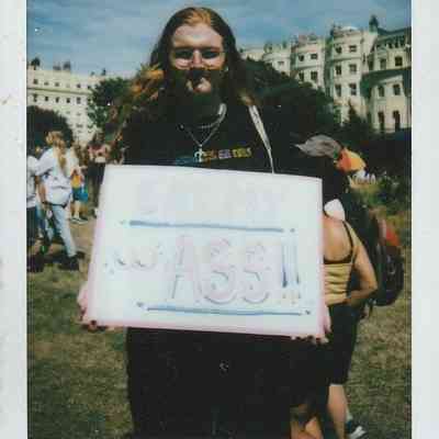 A person with long hair is holding a colourful sign which says 'EAT MY ASS".