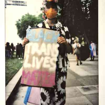 A person wearing a colorful mask and floral outfit holds a sign reading "Black Trans Lives Matter".