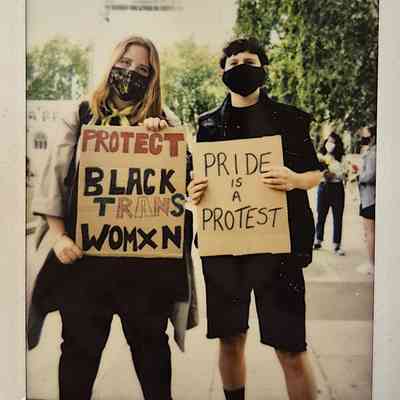 Two masked individuals hold signs which say "PROTECT BLACK TRANS WOMXN" and "PRIDE IS A PROTEST".