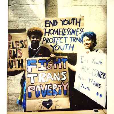 Two people hold signs advocating for ending youth homelessness and supporting trans youth.
