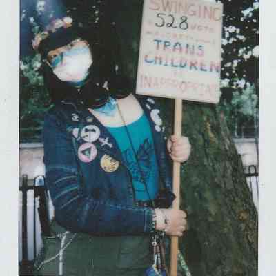 A masked person stands holding a sign expressing support for trans children under a tree.