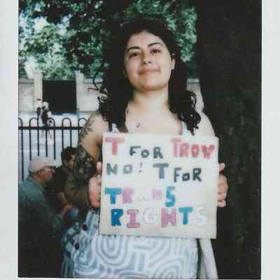 A person stands under a tree holding a sign advocating for trans rights in colorful letters.