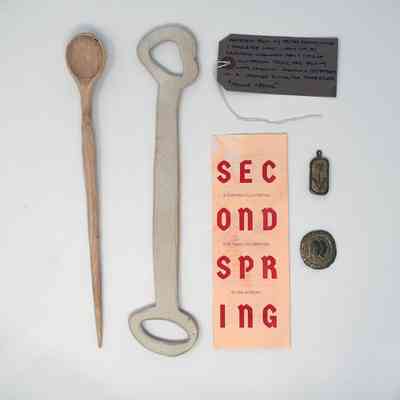 The image features a wooden spoon, bottle opener, note, pamphlet, and two small medals.