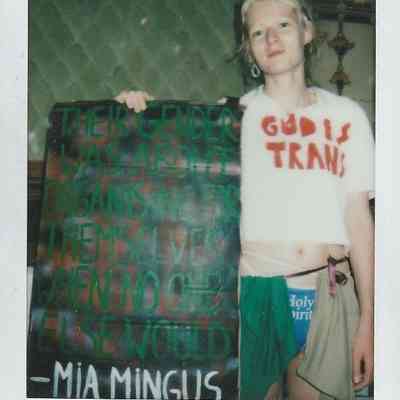 A person stands holding a colorful sign with a quote, wearing a shirt with "God is Trans."