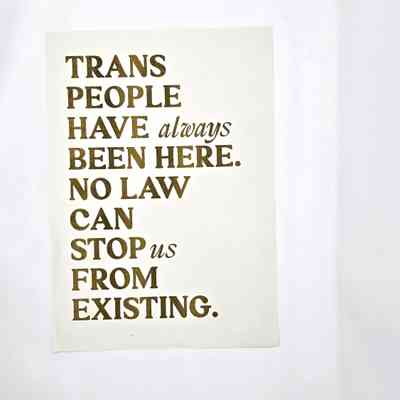 A print which has a white background and gold lettering which says 'Trans People Have Always Been Here. No Law Can Stop Us From Existing'
