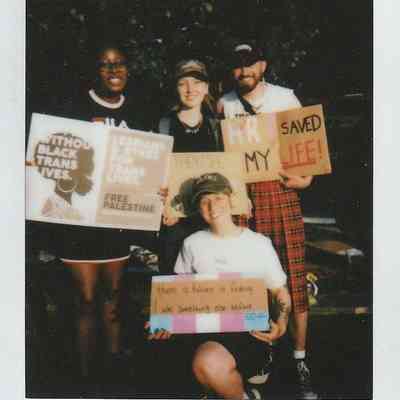 Four people are posing together outdoors, holding various protest signs that express support for trans rights.