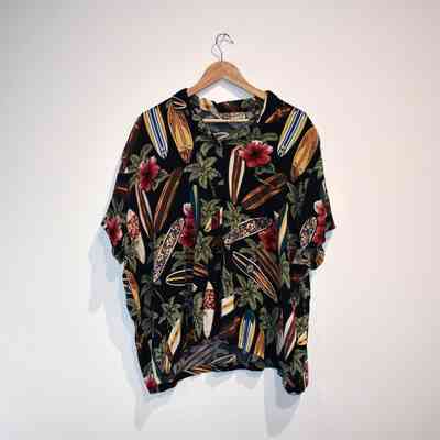 A colourful Hawaiian shirt featuring surf boards, green leaves and red flowers on a back background, on a wooden hanger.