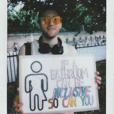 A person holds a sign saying, "If a bathroom can be inclusive, so can you," outdoors.