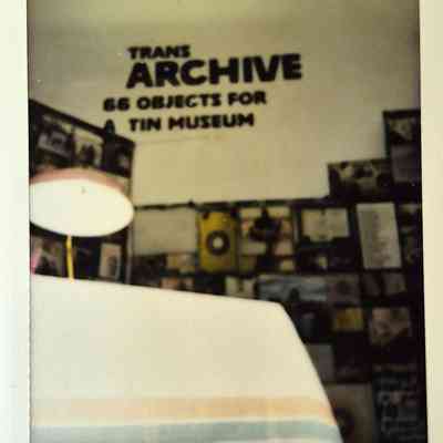A room displays a sign reading "TRANS ARCHIVE: 66 Objects For A Tin Museum".
