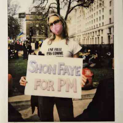 A person stands outdoors holding a sign reading "Shon Faye for PM," wearing a pink mask.