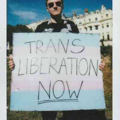 A person holds a sign saying "Trans Liberation Now," standing outside near a white building.
