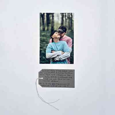 A photo of two people tenderly embracing in a wood celebrating their engagement. Both wear baseball caps, one a blue t-shirt, the other a pink t-shirt. With a brown handwritten luggage tag.