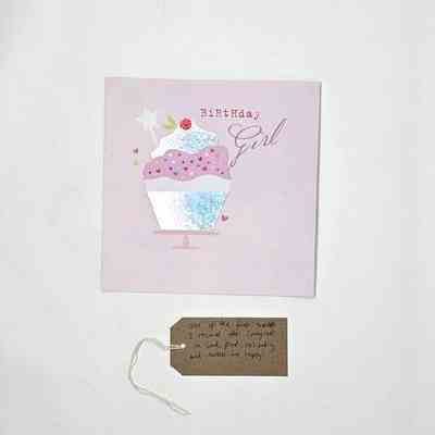 A Birthday Card which says 'Birthday Girl' and has an illustration of cupcake. The card is predominantly pink colours. Underneath the greeting card is a brown tag with writing on it.