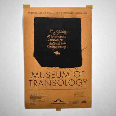 Beige poster reading "Brighton Museum & Art Gallery / extended to October 2019" [image of black fabric with embroidery reading "My gender & transness cannot be defined in a single image."] "MUSEUM OF TRANSOLOGY". under this there are some credits listed along with logos and other information.