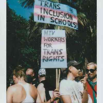 A group of people hold signs, one which says "TRANS INCLUSION IN SCHOOLS" and the other "HIV WORKERS FOR TRANS RIGHTS".