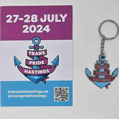 A blue, white and pink flyer for Trans Pride Hastings 2024 and an enameled metal keyring in the shape of the Trans Pride Hastings logo. The logo is a blue anchor with a pink heart a the top and the words "Trans Pride Hastings" in white on a pink ribbon entwined round the anchor.