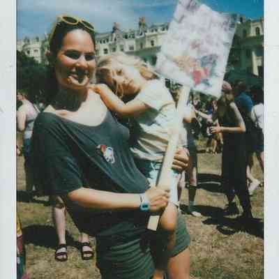 A smiling woman holds a sign with a child at an outdoor gathering on a sunny day.